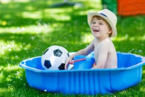 Young boy sitting in a blue inflatable pool with a soccer ball and toy shovel, enjoying summer play.