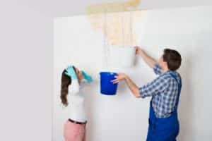 Couple inspecting a water leak in a home, with one person holding a bucket under the leak and the other looking concerned, illustrating the importance of checking for plumbing issues.