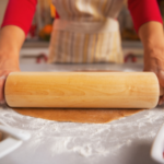 Hands rolling dough with a wooden rolling pin on a floured countertop, showcasing home cooking and preparation.