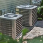 Two air conditioning units on a paved surface, surrounded by greenery, by The Meridian Company.