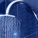 Chrome showerhead with water droplets on a blue tiled wall, by The Meridian Company.