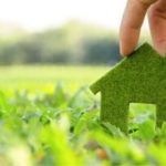 Hand holding a green house shape made of grass in a blurred outdoor setting, promoting eco-friendly home services.