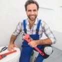 Smiling technician in blue overalls holds a red pipe wrench in a bright bathroom, representing The Meridian Company.