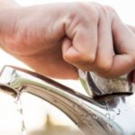 A hand turning on a shiny metallic faucet with water droplets, representing plumbing services by The Meridian Company.