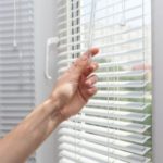 Hand reaching for partially open window blinds in a modern frame, allowing natural light to enhance home air quality.
