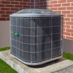 Air conditioning unit with metallic finish on concrete slab, surrounded by grass and a contrasting brick wall.
