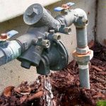 Water meter with pipes and valves on mulched ground by The Meridian Company, showcasing plumbing infrastructure.