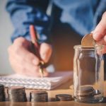 Hands writing in a notebook with coins and a jar, illustrating financial planning by The Meridian Company.