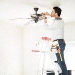 Man on ladder repairing ceiling fan, showcasing home maintenance by The Meridian Company in a bright interior setting.