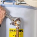 Person using an adjustable wrench on a water heater with copper pipes, part of The Meridian Company plumbing services.