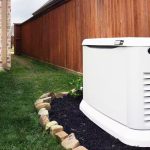 Generator unit in garden with stone border and wooden fence, showcasing home maintenance by The Meridian Company.