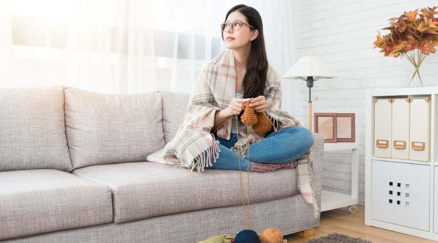 Young woman knitting on a gray couch surrounded by yarn and supplies, emphasizing cozy home services by The Meridian Company.
