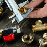 Hand holding a caliper measuring brass plumbing fittings on a dark wooden surface for The Meridian Company's services.