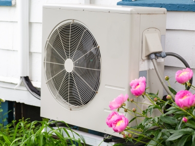 Outdoor condenser unit of a mini split heat pump system beside blooming pink peonies, illustrating ductless heating and cooling technology.
